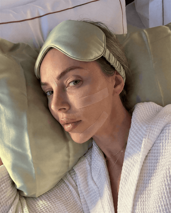 Kvinna ligger i sängen med silikontejp på ansiktet som del av hudvårdsrutin under natten.